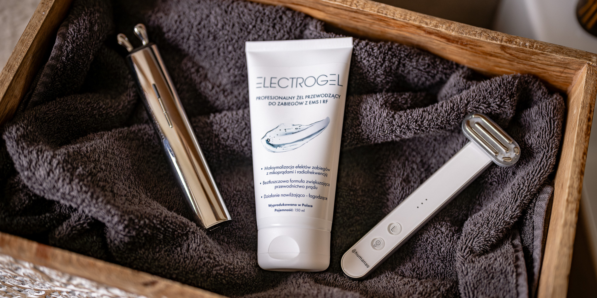 Żel przewodzący Humanas Electrogel - 150 ml umieszczony w drewnianej skrzyni na ręczniku obok urządzeń do pielęgnacji beauty