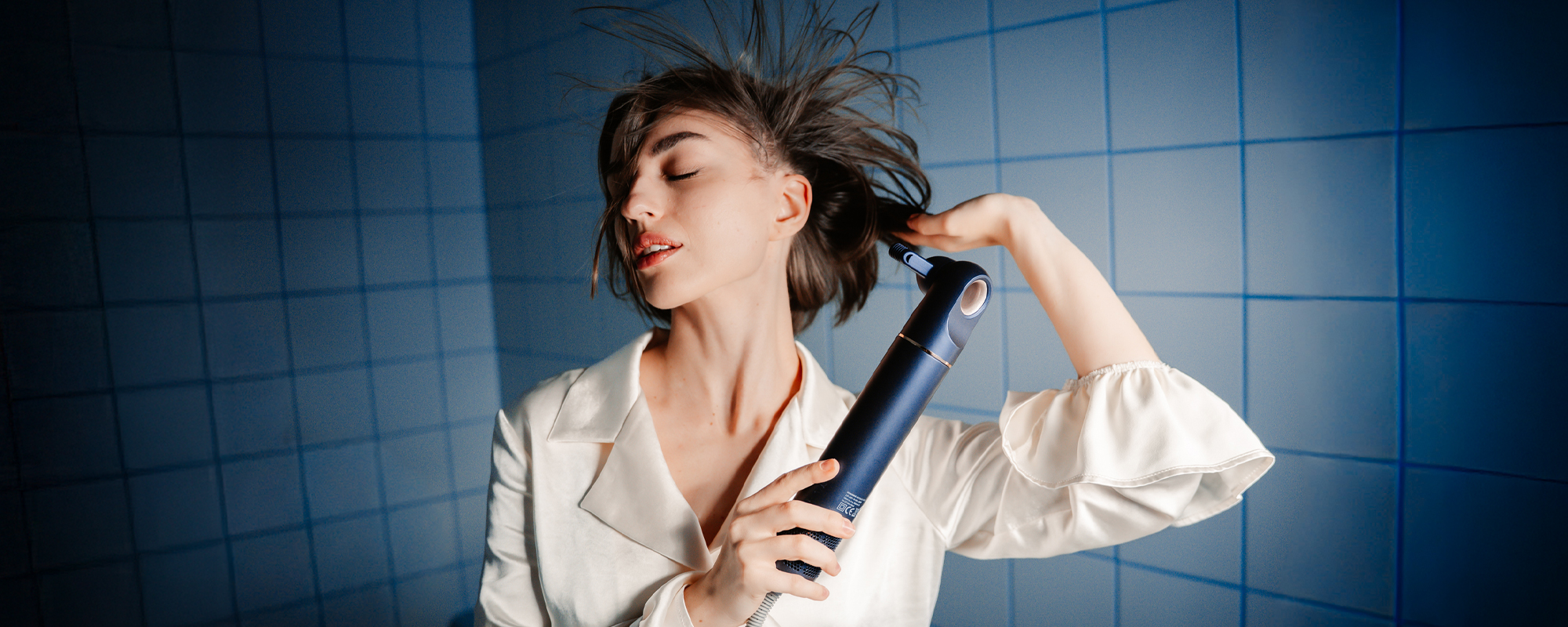 Kobieta suszy włosy za pomocą suszarki do włosów z funkcją jonizowania Humanas AirTouch HD-AT05. W tle kafelki łazienkowe.