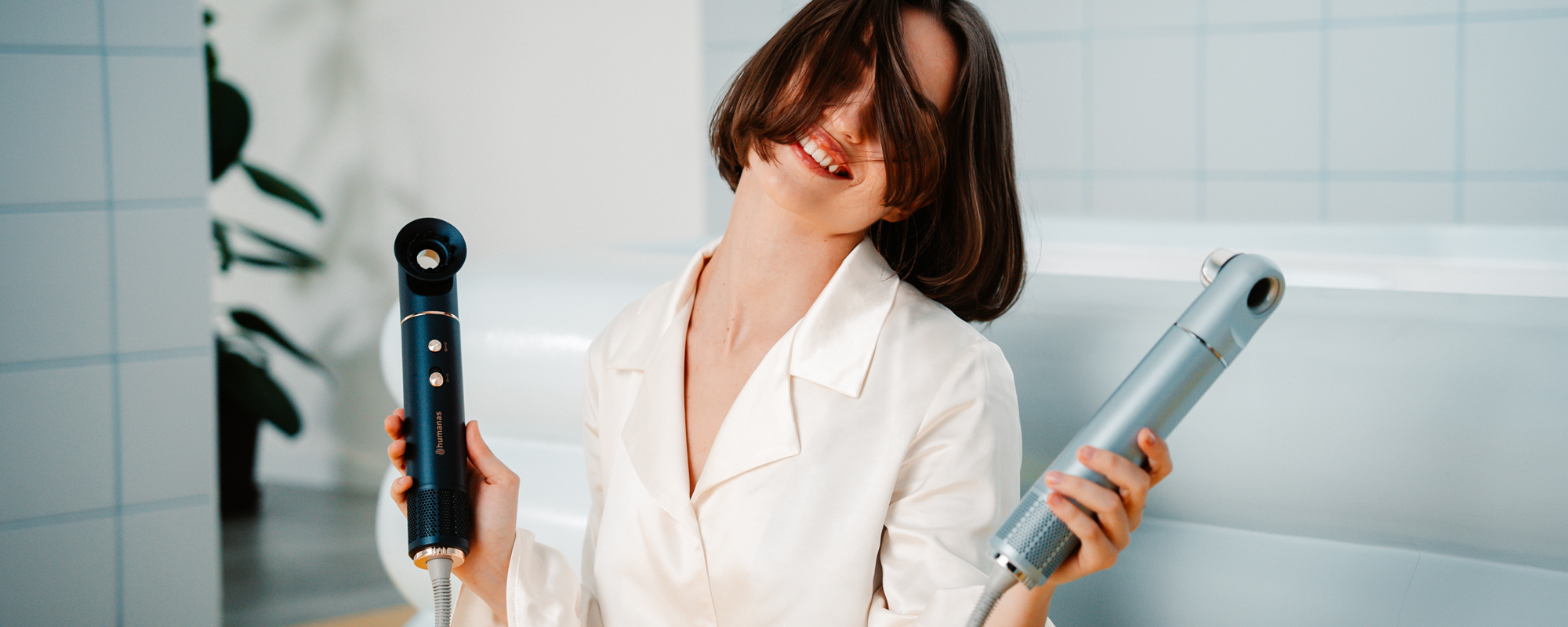 Kobieta trzymająca w dłoniach dwie suszarki do włosów z funkcją jonizowania Humanas AirTouch HD-AT05. W tle kafelki oraz wanna.