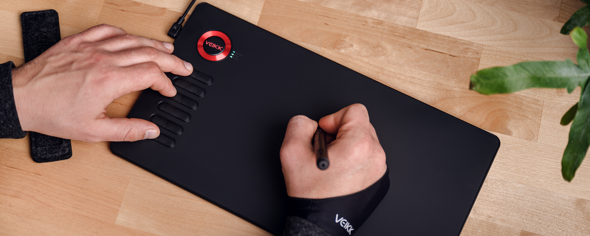Zdjęcie - tablet graficzny Veikk A15 na jasnobrązowym biurku, lewa dłoń mężczyzny naciska klawisz funkcyjny, prawa obsługuje piórko