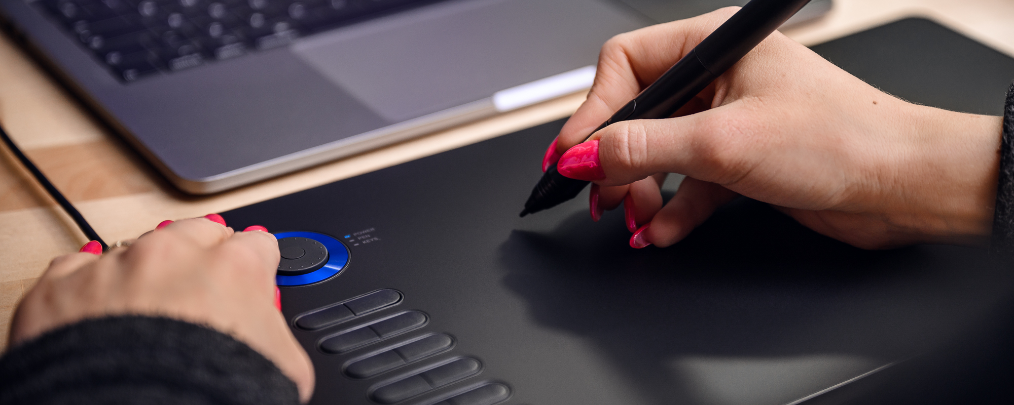Zdjęcie - tablet graficzny Veikk A15 Pro na jasnobrązowym biurku, lewa dłoń kobiety naciska klawisz funkcyjny, prawa obsługuje piórko pasywne Veikk